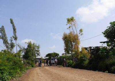 Photograph of several people standing on a dirt road outside a white van in the distance. Greenery and trees line the road on either side.